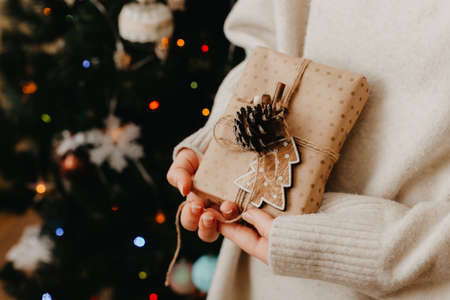 girl hands holding small gift in her hands. Close up. decorated Christmas tree on background. Bright Christmas card with space for text with space for textの写真素材