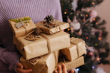 Closeup of woman in violet pullover holding many gift boxes in her hands. Decorated Christmas tree on background. Gift boxes wrapped in craft paperの写真素材
