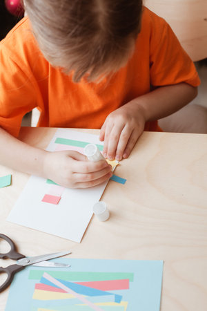 Cute little girl sits at desk and makes Christmas card. Creating Christmas tree from strips of...