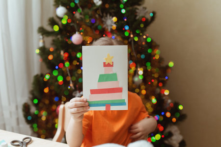 Cute little girl sits at desk and makes Christmas card. Creating Christmas tree from strips of...