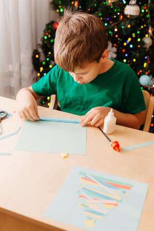 Cute little boy sits at desk and makes Christmas card. Creating Christmas tree from strips of colored paper.の写真素材