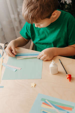 Cute little boy sits at desk and makes Christmas card. Creating Christmas tree from strips of colored paper.の写真素材