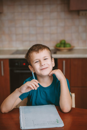 Cute child writing letters in notebook sitting by table in kitchenの写真素材
