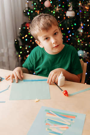 Cute little boy sits at desk and makes Christmas card. Creating Christmas tree from strips of colored paper.の写真素材