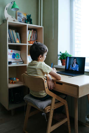 7 years old boy sitting by desk with laptop doing writing task during online lesson. Back view. Smiling woman tutor on computer screenの写真素材