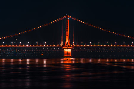 The Yangtze River Bridge in Wuhan illuminated in warm lights with colorful reflections on calm river...