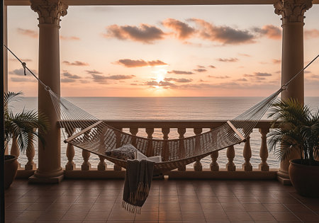 A woven hammock hangs on a balcony overlooking the ocean at sunset, complete with a pillow and blanket, framed by columns and potted plants.の素材