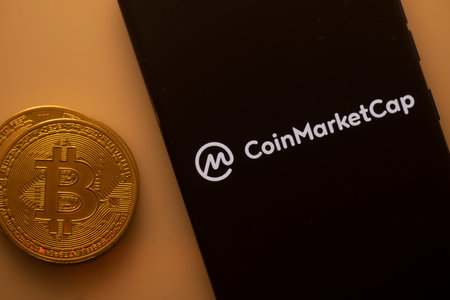 Dhaka, Bangladesh 22 September 2025:Bitcoin coin beside a phone showing the CoinMarketCap logo on a...