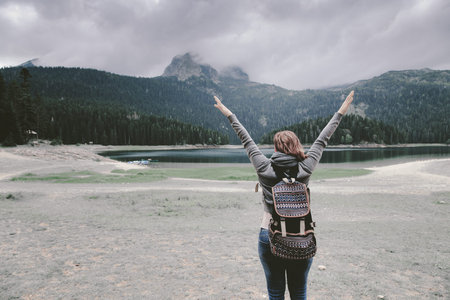 Woman with backpack standing near lake and mountain, hands upの写真素材