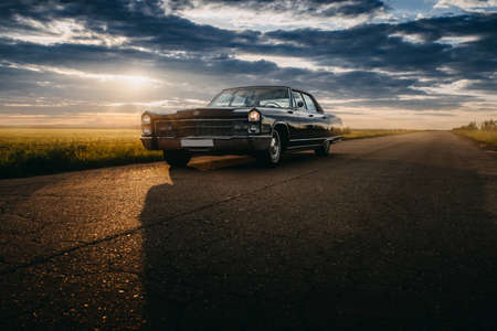 Engels, Russia - June 08, 2018: Black retro vintage muscle car Cadillac Fleetwood Brougham is parked at countryside asphalt road at golden sunset