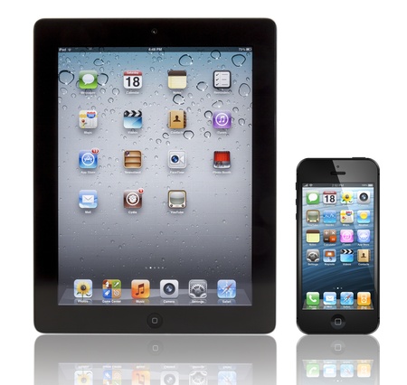 A black Apple iPhone 5 in front of a black Apple iPad 3 tablet. Both devices are showing the homepage with the same default wallpaper and are isolated on white on a reflective surface.のeditorial素材