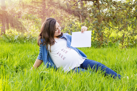 Beautiful pregnant woman outdoor with a paper in hand show the day birth of her babyの写真素材