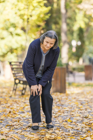 Senior woman walking in autumn park and having knee pain. Arthritis pain concept.の写真素材