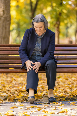 Senior woman walking in autumn park and having knee pain. Arthritis pain concept. Focus on her face.の写真素材