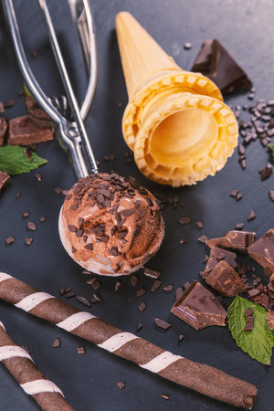 Chocolate ice cream in scoop with wafer sticks, cone and chocolate on a black slate board. Focus on ice cream in scoop.の写真素材