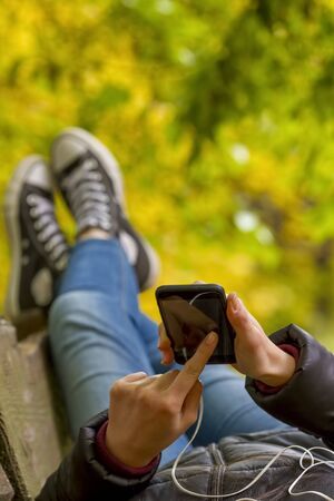 Teenage girl using smartphone lying on the bench in autumn park. Focus on smart phone.の写真素材