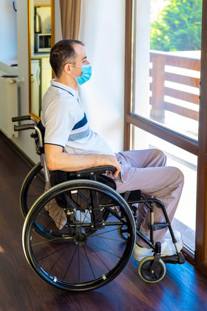 Smiling Young Handicapped Man Sitting On Wheelchair In Kitchen.  Young man wearing face mask sitting infront of kitchen. Focus on his face.の写真素材