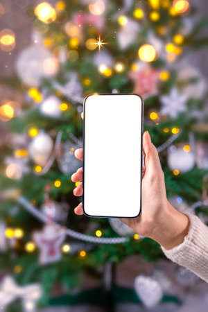 Close up smartphone with empty screen mockup on holiday background with Christmas tree, customer holding phone in hand, shopping online, purchasing gifts, searching information, browsing appsの写真素材