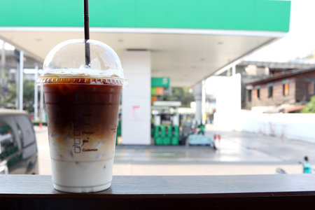 Ice coffee in plastic glass and straw on dark brown table and blur gas station background in Thailand.の写真素材
