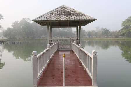Red bridge connect between white pavilion in mash and land ,behind is trees and reflect on water in swamp in the morning, a little fog cover above, Thailand.の写真素材