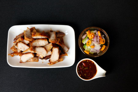 Grilled pork shoulder slice in white rectangle dish with hot and sour Thai style sauce in white cup and vegetable slice (chilli, cucumber, onion) in vinegar glass cup on black background.の写真素材