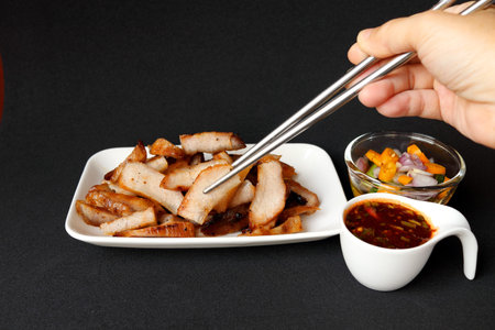 Right hand use chopsticks clamping grilled pork shoulder slice in white rectangle dish with hot and sour Thai style sauce in white cup and vegetable slice in vinegar glass cup on black background.の写真素材
