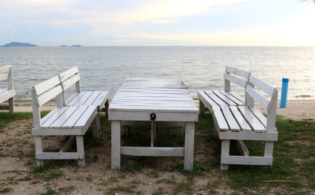 Set white painted table and benches on sand and sea, sky background with sunlight, white cloud in...