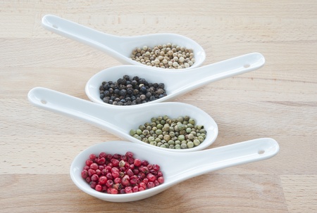 four kinds of peppercorns with white porcelain condiment spoons on a wooden tableの写真素材