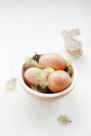 three brown chicken eggs on the feathers in a wooden bowl on a wooden table, close to the cut of birch bunnyの写真素材
