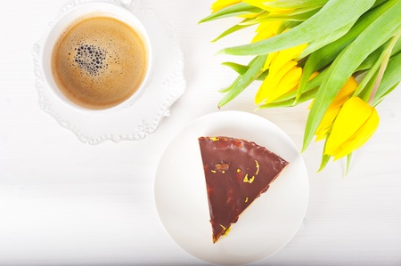 piece of layer cake with chocolate on a white plate, a cup of coffee and a bouquet of yellow tulipsの写真素材