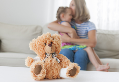 A teddy bear with a bandage paw. Mom kisses her daughter in the backgroundの写真素材