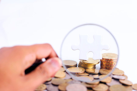 Businessman hand holding magnifying glass, white puzzle and gold coins with copyspace areaの写真素材