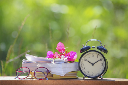 Black alarm clock stacked on old dictionary with pink flower and glasses.の写真素材