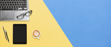 Vibrant office desk table working space with laptop, tablet, eyeglasses and a cup of hot coffee on yellow blue textures background with copy space, Flat lay for banner, wallpaper, business conceptの写真素材