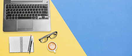 Modern desk office workplace with laptop, notebook, eyeglasses and a cup of hot coffee on yellow blue textures background with copy space, Flatlay for banner, wallpaper, business, education conceptの写真素材