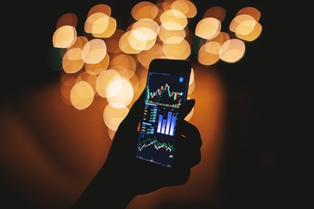 Silhouette of female hand holding smart phone in the dark with stock trading on display with light bokeh blurred background, Mobile business conceptの写真素材