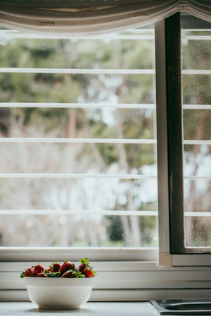 Strawberries in a white bowl with a window behind on a neutral background with trees in the...