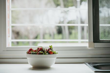 Strawberries in a white bowl with a window behind on a neutral background with trees in the distance.の写真素材