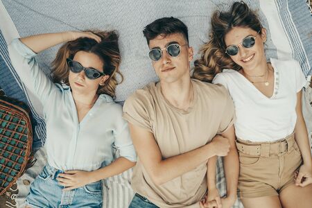 Young people sunbathing, wearing sunglasses, on a picnic blanket by a lake in summer.の写真素材