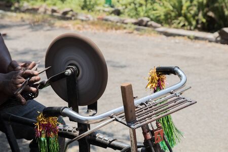 an Indian man holding a steel scissor in his hand to sharpen it on the rotating whetstone which is connected to the rear wheel of his bicycle through a stringの写真素材