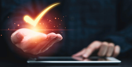 A hand emerges from a digital device, holding a bright, glowing checkmark symbolizing achievement and confirmation.の写真素材