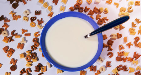 A top-down view of a blue bowl filled with creamy soup, with a dark spoon inserted. Alphabet pasta is scattered around the bowl.の写真素材
