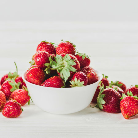A bright, close-up shot of a white bowl overflowing with vibrant red strawberries, with more scattered around.の写真素材