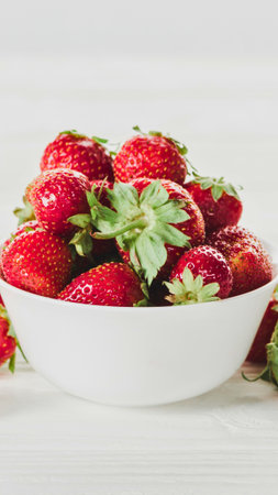 A close-up shot of a white bowl overflowing with bright red, ripe strawberries, their green leaves adding a vibrant contrast.の写真素材