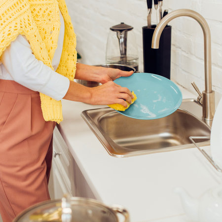 A person is actively cleaning a blue plate with a yellow sponge in a stainless steel kitchen sink. The faucet is on.の写真素材