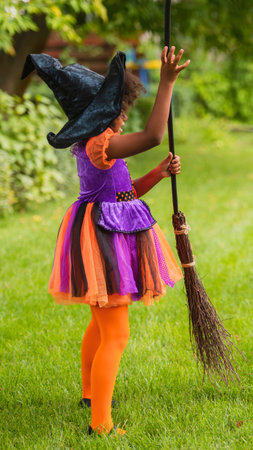 A child dressed as a witch, wearing a black hat and colorful tutu, holds a broomstick in a grassy outdoor setting.の写真素材