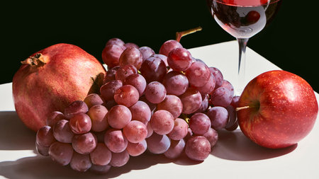 A still life arrangement featuring a pomegranate, red grapes, and an apple next to a glass of red wine.の写真素材