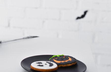 Two decorated Halloween cookies, one ghost and one jack-o'-lantern, sit on a black plate against a white brick wall.の写真素材