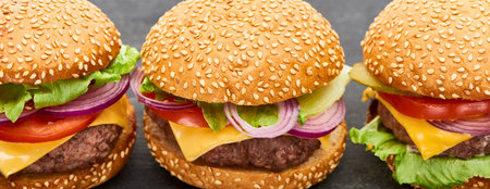 A close-up, eye-level view of three freshly made hamburgers with sesame seed buns, cheese, lettuce, tomato, and red onion.の写真素材
