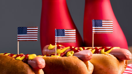 A close-up of several hot dogs adorned with small American flags and mustard, with red ketchup bottles in the background.の写真素材